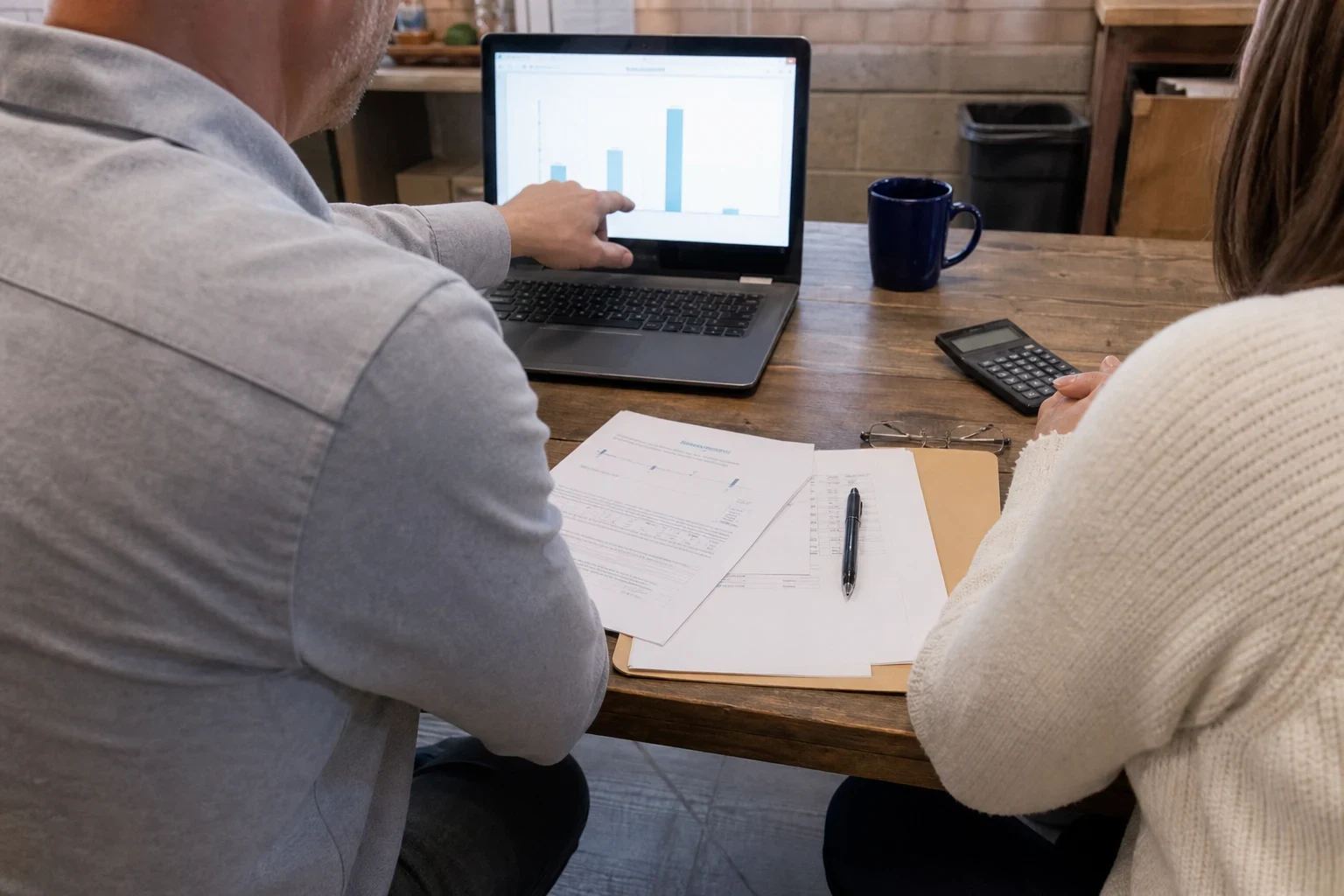Man in a gray button-down points to a bar chart on a laptop while a woman in a cream sweater looks on at a wooden table with papers, a pen, a calculator and a mug