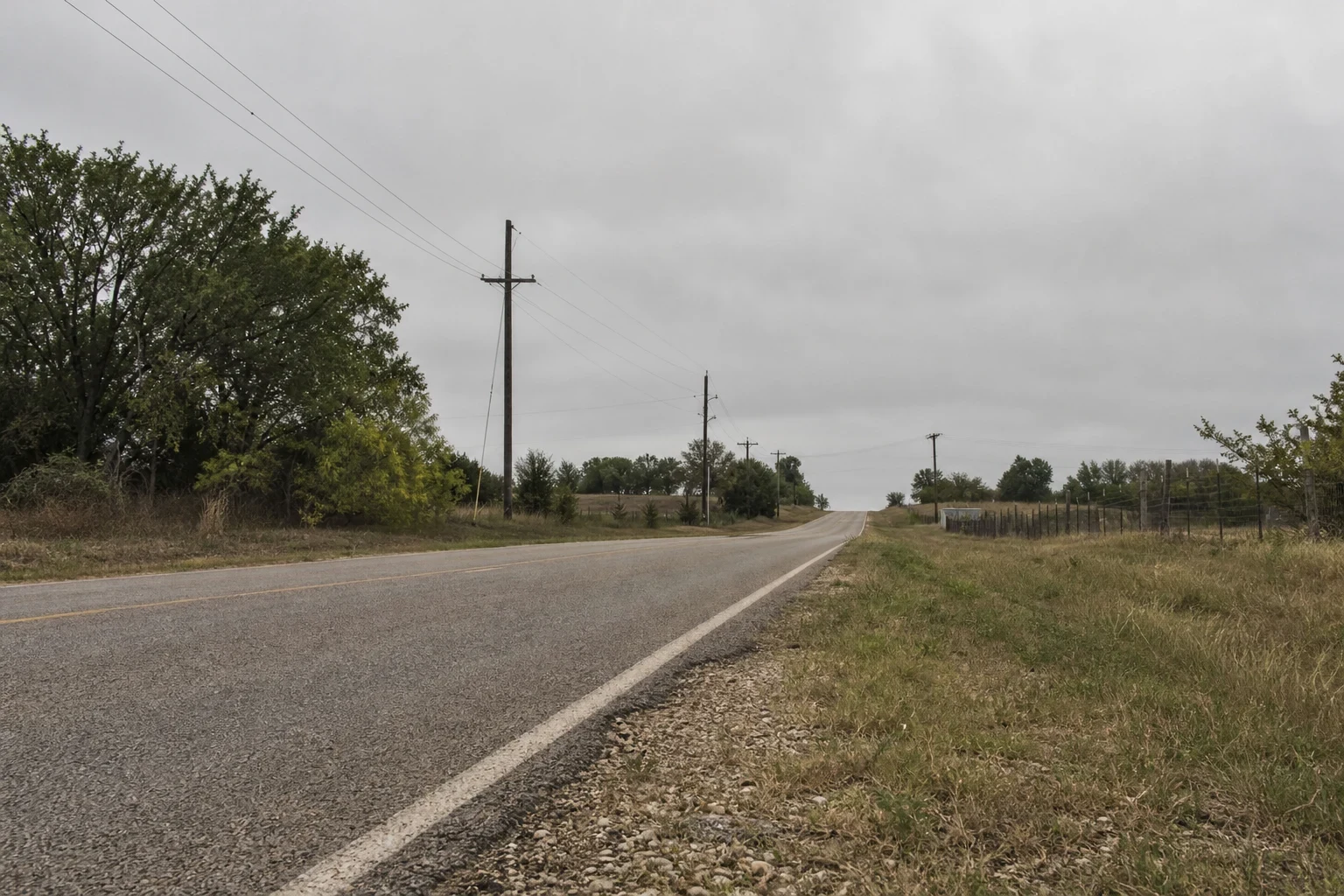 Quiet two-lane rural road under an overcast sky, with utility poles, trees, a wire fence and grassy shoulders on the edge of town