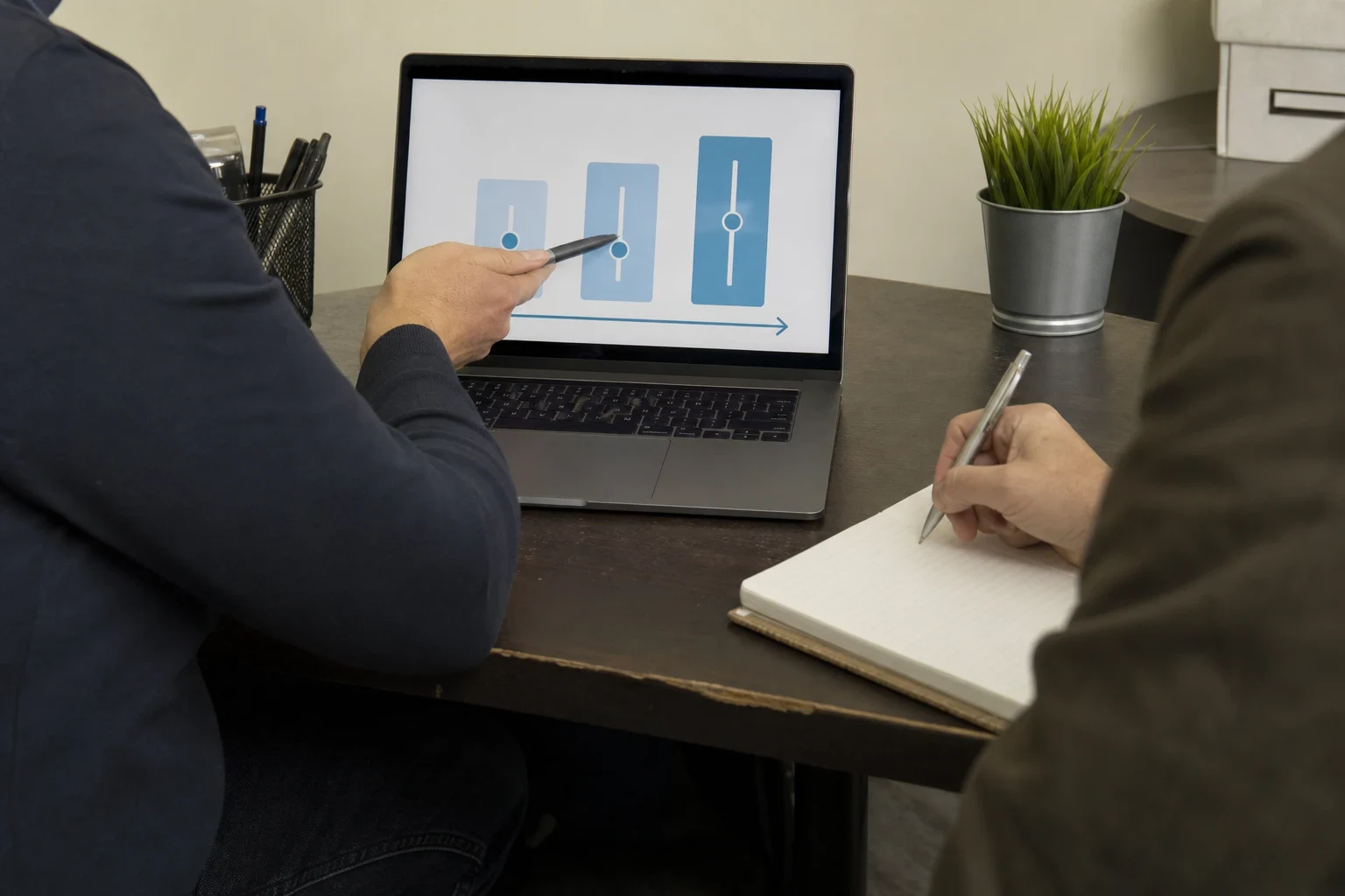Agent points a pen at a laptop showing three adjustable slider bars while a client takes notes in a notebook at a worn office desk with a pen cup and small plant