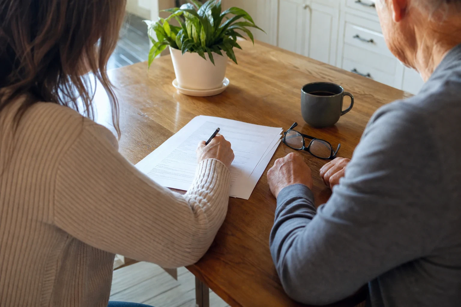 A woman in a beige cardigan writes on a short paper form at a wooden kitchen table beside an older man, with reading glasses, a coffee mug and a potted plant nearby